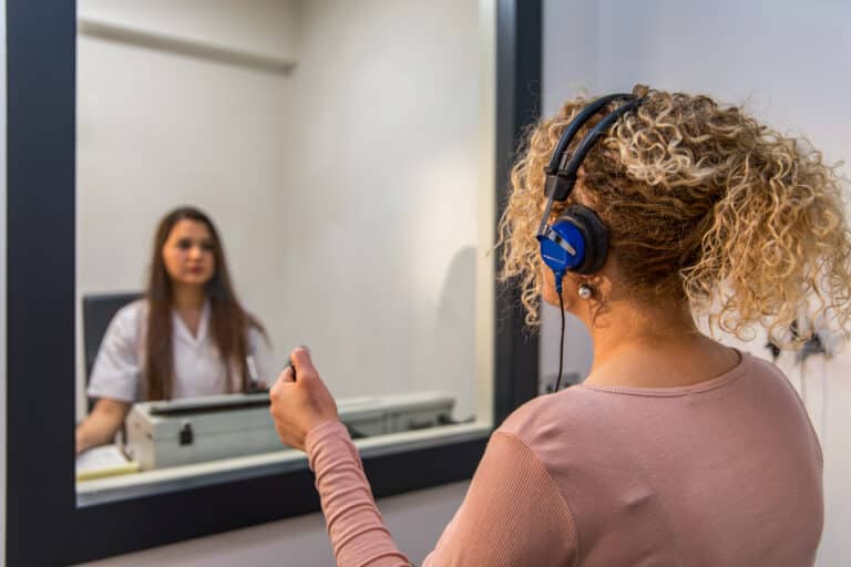 A younger woman takes a hearing test in an audiologist's sound booth.