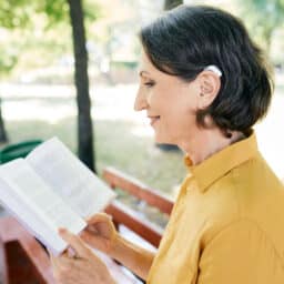 Woman wearing a hearing aid while reading a book in a public park.