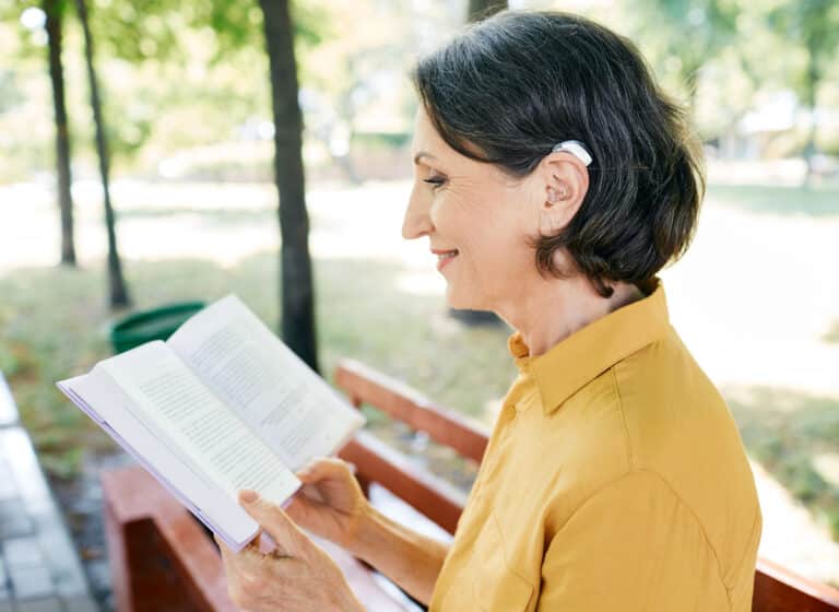 Woman wearing a hearing aid while reading a book in a public park.